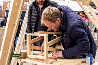 Image of a man at an outdoor workshop, bent over building a wood chair