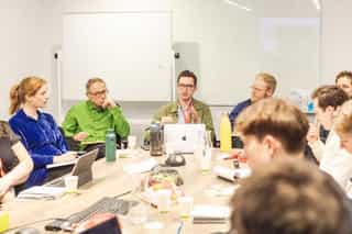 An image of people gathered around a table one man talks as the others listen intently