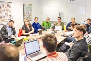 An image of people gathered around a table one man talks as the others listen intently