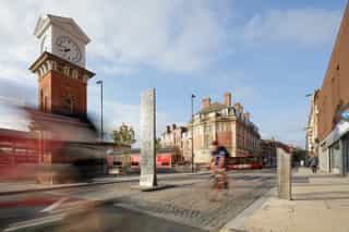 Road improvements outside Altrincham bus interchange, with a new gateway art piece in the middle of the road and the historic clock tower in the distance. A cyclist is passing through the street.