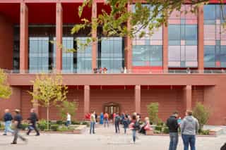 An image of people outside Anfield, a few are on the balcony above, a few are walking past, a couple are sat on the benches admiring the planting, and some people are gathered looking at the Plaque on the wall