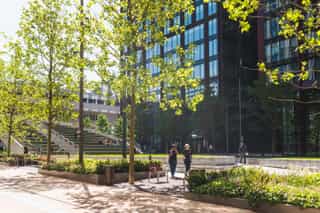Image showing a city centre park. Terraced green steps and a lawn area with flower beds is surrounded by high rise glass buildings. Two people are walking through the space.