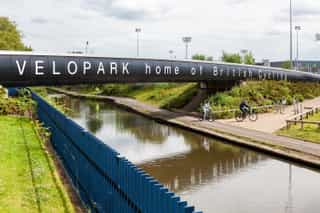 An image of a bridge over a canal with the words 'VELOPARK home of British Cycling' on the side, two people are cycling down the path on the canal