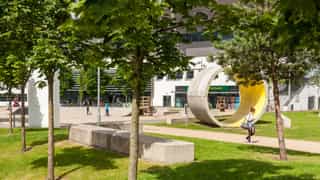 An image of a small grassland with trees and concrete slabs for seating, a concrete cylinder is placed upright on the ground,. A woman walks up a path in the middle while kids use scooters in the background