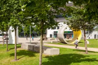 An image of a small grassland with trees and concrete slabs for seating, a concrete cylinder is placed upright on the ground,. A woman walks up a path in the middle while kids use scooters in the background