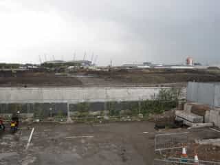 A before image of Velopark - a grey scene of the start of construction, a cloudy sky, concrete wall, metal fencing and diggers digging the ground