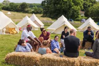Photo showing people sat in a field on hay bales in a discussion, with bell tents pitched in the background.
