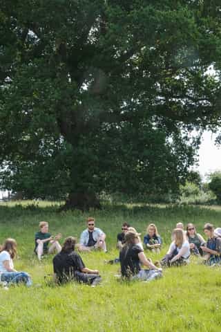 An image of people gathered sitting in a circle on grass with a large tree behind them