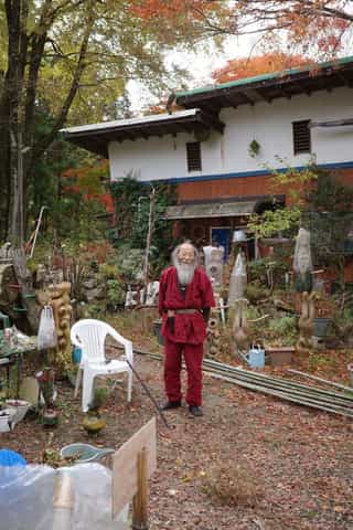 An full length image of a bearded man in a red Samue smiling towards the camera. In front of a house, plants, old sculptures, pots and a chair are scattered around him.
