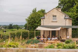 A long shot image of a building with a canopy in the countryside, people are sat gathered round a table underneath the canopy