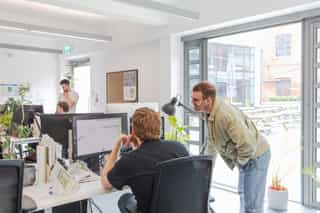 An image of a man sat at his computer with another man standing over his shoulder reviewing his work