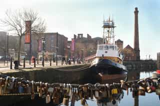 An image of a boat at the Liverpool Dock, people walk past the boat. In the bottom of the image, a railing filled with locks attached to it.