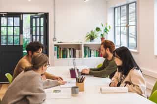 An image of four people gathered round a desk working, they have books, markers and a laptop on the table