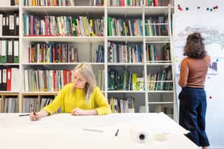 An image of a woman sat writing, with a wall of books behind her