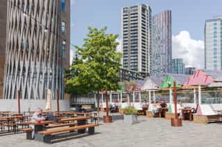 An exterior shot of Sugar House Island, people are using outdoor seating, bench table, some of which have a canopy roof for shade, towering buildings are in the distance