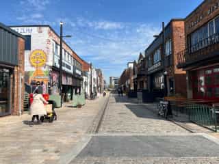 An image looking down a street in Hull, a woman pushes her child in a pram, canopy lighting guides you down the street, there is colourful street art on the wall, it is a bright sunny day